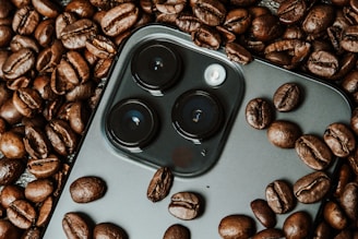 A smartphone camera module is surrounded by roasted coffee beans. The camera lenses are prominently featured among the beans, providing a contrast between technology and organic elements.