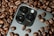 A smartphone camera module is surrounded by roasted coffee beans. The camera lenses are prominently featured among the beans, providing a contrast between technology and organic elements.