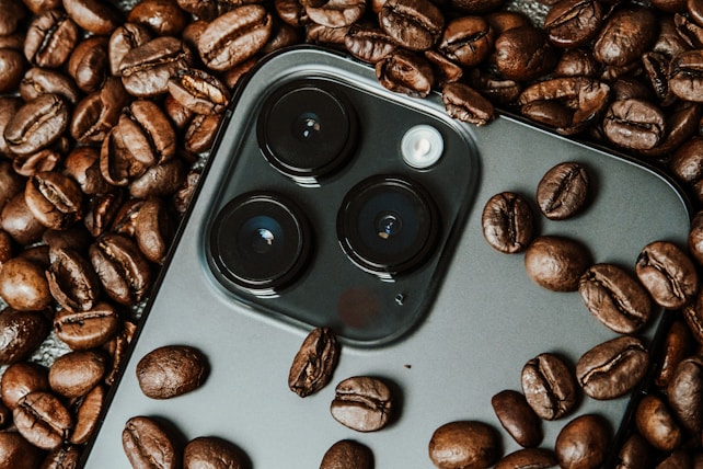 A smartphone camera module is surrounded by roasted coffee beans. The camera lenses are prominently featured among the beans, providing a contrast between technology and organic elements.