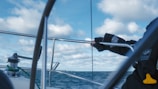 A person wearing dark gloves is gripping the rail of a sailboat, with the ocean visible in the background under a partly cloudy sky. The scene conveys a sense of being out at sea, with metal poles and sailing equipment in view.