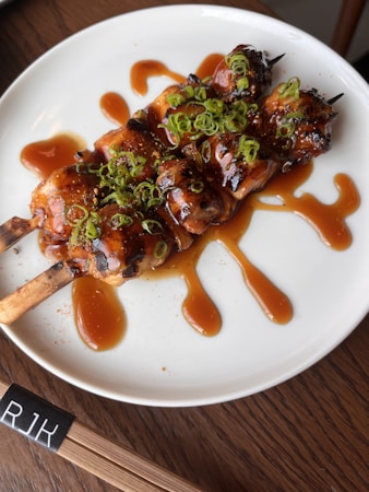 Two grilled skewers topped with green onions are drizzled with a glossy brown sauce, presented on a white plate. The skewers and sauce create an appetizing display, accompanied by chopsticks with the initials 