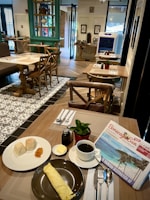 A cozy cafe setting with wooden tables and chairs, featuring a plate with two bread rolls and jam, a large rolled omelette on a separate plate, a small dish of butter, a potted plant, and a cup of coffee next to a newspaper. The interior has tiled and wooden floors, decorative elements, and natural light coming through the large windows.