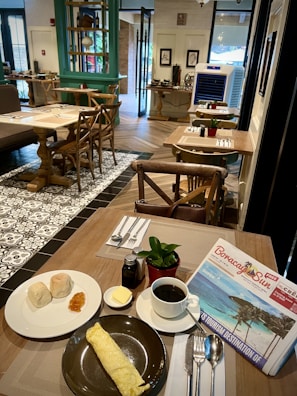 A cozy cafe setting with wooden tables and chairs, featuring a plate with two bread rolls and jam, a large rolled omelette on a separate plate, a small dish of butter, a potted plant, and a cup of coffee next to a newspaper. The interior has tiled and wooden floors, decorative elements, and natural light coming through the large windows.