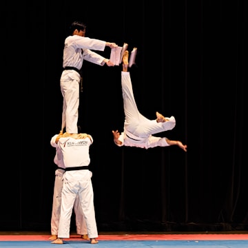 Four martial artists perform an acrobatic move. Two individuals stand side by side holding one person on their shoulders, while another performer flips in mid-air attempting to break boards held by the elevated individual.