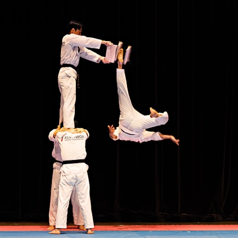 Four martial artists perform an acrobatic move. Two individuals stand side by side holding one person on their shoulders, while another performer flips in mid-air attempting to break boards held by the elevated individual.