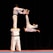 Four martial artists perform an acrobatic move. Two individuals stand side by side holding one person on their shoulders, while another performer flips in mid-air attempting to break boards held by the elevated individual.