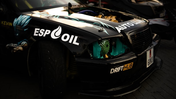 A customized black car with colorful decals and motifs, including an anime character on the side. The car features various decals such as 'ESP OIL,' 'AUTO LAB,' and 'DRIFTHUB.' The license plate reads 'GOKU' and part of the engine is visible, showing elements of gold and turquoise. The car is parked on a paved surface.