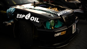 A customized black car with colorful decals and motifs, including an anime character on the side. The car features various decals such as 'ESP OIL,' 'AUTO LAB,' and 'DRIFTHUB.' The license plate reads 'GOKU' and part of the engine is visible, showing elements of gold and turquoise. The car is parked on a paved surface.