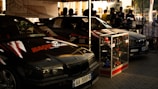 Two black racing cars with logos and decals are parked side by side under a canopy. A display case filled with various car parts stands in front of one car. Several people are gathered in the background, possibly discussing or observing the cars.