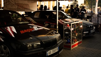 Two black racing cars with logos and decals are parked side by side under a canopy. A display case filled with various car parts stands in front of one car. Several people are gathered in the background, possibly discussing or observing the cars.