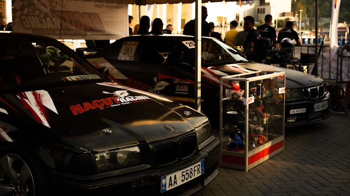 Two black racing cars with logos and decals are parked side by side under a canopy. A display case filled with various car parts stands in front of one car. Several people are gathered in the background, possibly discussing or observing the cars.