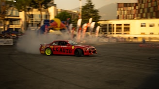 A red sports car is performing a drift maneuver on an outdoor track surrounded by buildings. Smoke emerges from the tires, indicating high speed and skillful driving. Brightly colored banners and flags can be seen in the background, suggesting a racing event or competition. The sun casts a warm glow over the entire scene.
