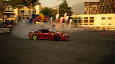 A red sports car is performing a drift maneuver on an outdoor track surrounded by buildings. Smoke emerges from the tires, indicating high speed and skillful driving. Brightly colored banners and flags can be seen in the background, suggesting a racing event or competition. The sun casts a warm glow over the entire scene.
