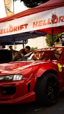 A red sports car with logos and racing decals is parked under a red tent. Part of the car's front and side is visible with black rims and a visible racing number '8'. There are people in the background and various banners and equipment suggesting a car event or race meet.