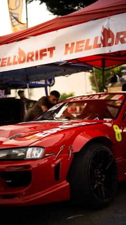 A red sports car with logos and racing decals is parked under a red tent. Part of the car's front and side is visible with black rims and a visible racing number '8'. There are people in the background and various banners and equipment suggesting a car event or race meet.