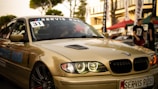 A sleek, beige BMW car is captured in what appears to be an outdoor automotive event. The car's headlights are illuminated, and a 'Servis Rusi' sticker is visible on the windshield. The background includes blurred figures and tents, suggesting a lively atmosphere.