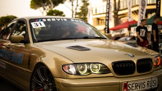 A sleek, beige BMW car is captured in what appears to be an outdoor automotive event. The car's headlights are illuminated, and a 'Servis Rusi' sticker is visible on the windshield. The background includes blurred figures and tents, suggesting a lively atmosphere.