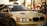 A sleek, beige BMW car is captured in what appears to be an outdoor automotive event. The car's headlights are illuminated, and a 'Servis Rusi' sticker is visible on the windshield. The background includes blurred figures and tents, suggesting a lively atmosphere.