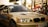 A sleek, beige BMW car is captured in what appears to be an outdoor automotive event. The car's headlights are illuminated, and a 'Servis Rusi' sticker is visible on the windshield. The background includes blurred figures and tents, suggesting a lively atmosphere.