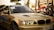 A sleek, beige BMW car is captured in what appears to be an outdoor automotive event. The car's headlights are illuminated, and a 'Servis Rusi' sticker is visible on the windshield. The background includes blurred figures and tents, suggesting a lively atmosphere.