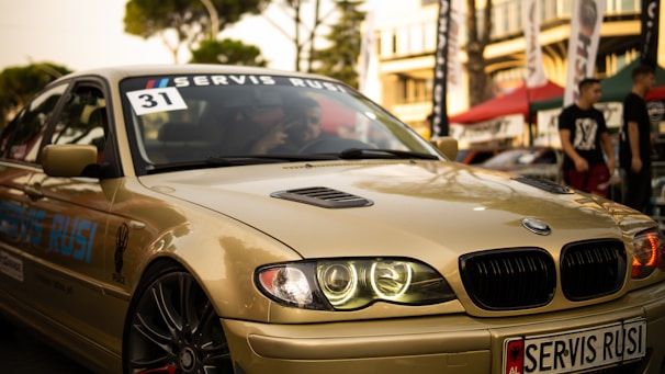 A sleek, beige BMW car is captured in what appears to be an outdoor automotive event. The car's headlights are illuminated, and a 'Servis Rusi' sticker is visible on the windshield. The background includes blurred figures and tents, suggesting a lively atmosphere.