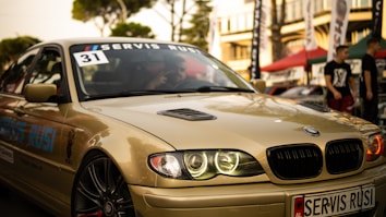 A sleek, beige BMW car is captured in what appears to be an outdoor automotive event. The car's headlights are illuminated, and a 'Servis Rusi' sticker is visible on the windshield. The background includes blurred figures and tents, suggesting a lively atmosphere.