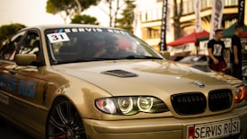 A sleek, beige BMW car is captured in what appears to be an outdoor automotive event. The car's headlights are illuminated, and a 'Servis Rusi' sticker is visible on the windshield. The background includes blurred figures and tents, suggesting a lively atmosphere.