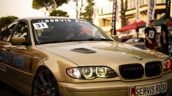 A sleek, beige BMW car is captured in what appears to be an outdoor automotive event. The car's headlights are illuminated, and a 'Servis Rusi' sticker is visible on the windshield. The background includes blurred figures and tents, suggesting a lively atmosphere.