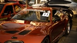 A shiny, modified Toyota Supra is parked in a lot with other cars. The car has a glossy red finish with aftermarket parts such as a large rear spoiler. The windshield displays the words 'Auto Servis Klodi' and 'Toyota Supra' along with a racing number sticker '11'. There are additional vehicles in the background, including an orange car with graphics and a silver one.
