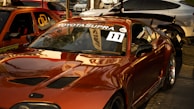 A shiny, modified Toyota Supra is parked in a lot with other cars. The car has a glossy red finish with aftermarket parts such as a large rear spoiler. The windshield displays the words 'Auto Servis Klodi' and 'Toyota Supra' along with a racing number sticker '11'. There are additional vehicles in the background, including an orange car with graphics and a silver one.