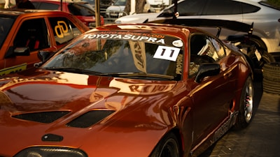 A shiny, modified Toyota Supra is parked in a lot with other cars. The car has a glossy red finish with aftermarket parts such as a large rear spoiler. The windshield displays the words 'Auto Servis Klodi' and 'Toyota Supra' along with a racing number sticker '11'. There are additional vehicles in the background, including an orange car with graphics and a silver one.
