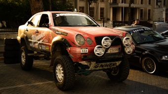A modified off-road vehicle with large tires and suspension sits prominently in a street setting. The vehicle is adorned with various sponsor decals, including 'Milwaukee', and features additional lights on the front. In the background, there are other parked cars and a building with tall windows and columns. The lighting suggests either early morning or evening.