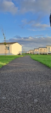A straight, paved pathway leading through a well-kept grassy area with mobile homes on either side. The sun casts shadows across the path, and the sky is partly cloudy with patches of blue.