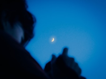 A crescent moon glows softly against a clear blue evening sky. In the foreground, the silhouette of a person appears slightly blurred, capturing a sense of quiet contemplation.