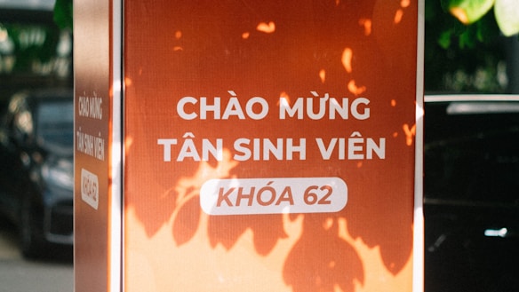 A vertical orange sign with white Vietnamese text welcoming new students, surrounded by natural light and shadows from leaves. The text in focus reads 'CHÀO MỪNG TÂN SINH VIÊN KHÓA 62.' A section of an automobile is faintly visible in the background.