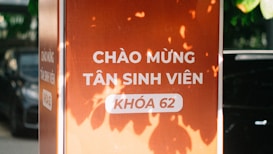 A vertical orange sign with white Vietnamese text welcoming new students, surrounded by natural light and shadows from leaves. The text in focus reads 'CH&Agrave;O MỪNG T&Acirc;N SINH VI&Ecirc;N KH&Oacute;A 62.' A section of an automobile is faintly visible in the background.