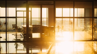 Sunlight streams into a room with large glass windows, casting reflections on the shiny floor. Several office chairs and a table are positioned near the windows, creating an airy and open atmosphere. The warm glow suggests evening light filling the space.
