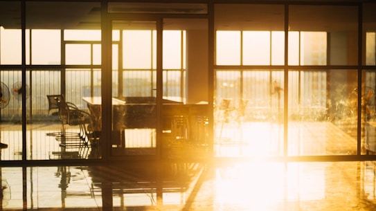 Sunlight streams into a room with large glass windows, casting reflections on the shiny floor. Several office chairs and a table are positioned near the windows, creating an airy and open atmosphere. The warm glow suggests evening light filling the space.