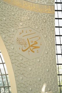 Intricate Islamic geometric patterns decorate a wall with Arabic calligraphy prominently displayed in gold. Sunlight filters through a series of grid-like window panes, casting soft shadows on the design.