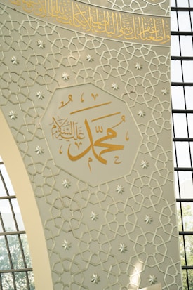 Intricate Islamic geometric patterns decorate a wall with Arabic calligraphy prominently displayed in gold. Sunlight filters through a series of grid-like window panes, casting soft shadows on the design.