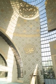 An intricately designed interior wall features elegant geometric patterns and Arabic calligraphy. The architecture includes large, arched windows allowing sunlight to cast detailed shadows across the decor. Dominant colors are light beige and gold, giving the space a serene and spiritual atmosphere.