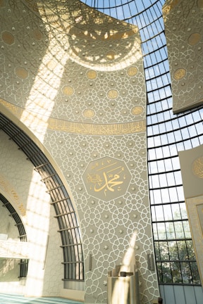An intricately designed interior wall features elegant geometric patterns and Arabic calligraphy. The architecture includes large, arched windows allowing sunlight to cast detailed shadows across the decor. Dominant colors are light beige and gold, giving the space a serene and spiritual atmosphere.