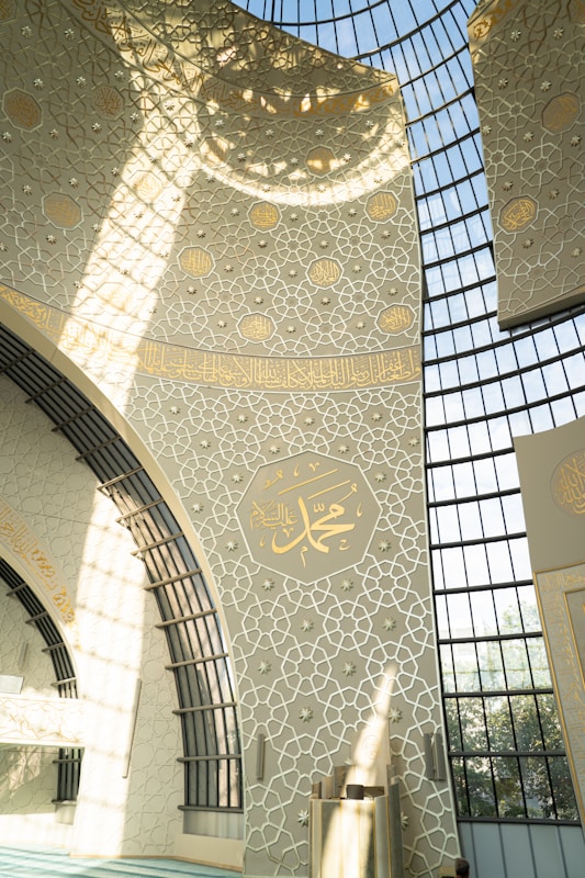 An intricately designed interior wall features elegant geometric patterns and Arabic calligraphy. The architecture includes large, arched windows allowing sunlight to cast detailed shadows across the decor. Dominant colors are light beige and gold, giving the space a serene and spiritual atmosphere.