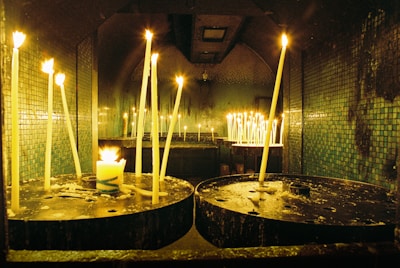 Tall, thin candles are lit and placed on circular metal holders in a dimly lit room with tiled walls. The flickering flames cast a warm glow, and there are melted wax drips on the holders.