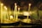 Tall, thin candles are lit and placed on circular metal holders in a dimly lit room with tiled walls. The flickering flames cast a warm glow, and there are melted wax drips on the holders.