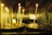 Tall, thin candles are lit and placed on circular metal holders in a dimly lit room with tiled walls. The flickering flames cast a warm glow, and there are melted wax drips on the holders.