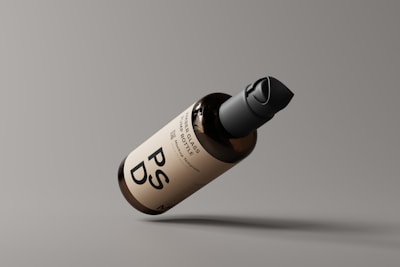 A brown glass pump bottle with a black cap is positioned diagonally against a simple gray background. The bottle features a minimalist label with bold black lettering, providing a clean and modern appearance.