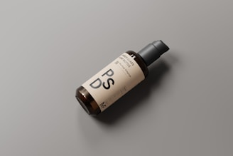 A dark brown glass bottle with a pump dispenser lies on a matte gray surface. The bottle is labeled with large letters 'PSD' and smaller text at the top and bottom. The label is a light beige color.
