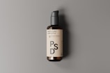 A dark amber glass pump bottle is positioned against a neutral gray background. The bottle features a cream-colored label with bold black text that reads 'PSD'. The pump on top is a matte black color, and the shadow cast suggests a light source from the left.