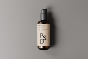 A dark amber glass pump bottle is positioned against a neutral gray background. The bottle features a cream-colored label with bold black text that reads 'PSD'. The pump on top is a matte black color, and the shadow cast suggests a light source from the left.